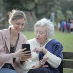 Woman showing an elderly grandma something on her smartphone while sitting on a bench