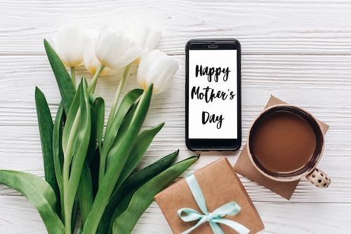 for unforgettable Mother's Day gifts blog post | happy mothers day text sign on stylish gift and phone and tulips and coffee on white wooden rustic background. flat lay with flowers and gadget with empty screen with space for text.