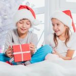 Christmas gifts for brother blog post - two kids wearing Santa hats and pajamas and sharing Christmas gift boxes in Christmas designed atmosphere